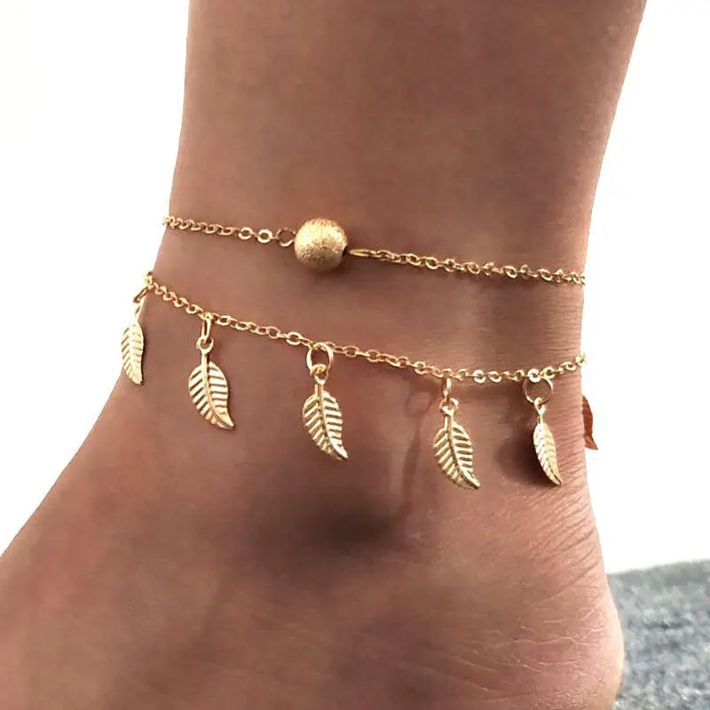 Gold anklets with leaf charms on a foot against a neutral background