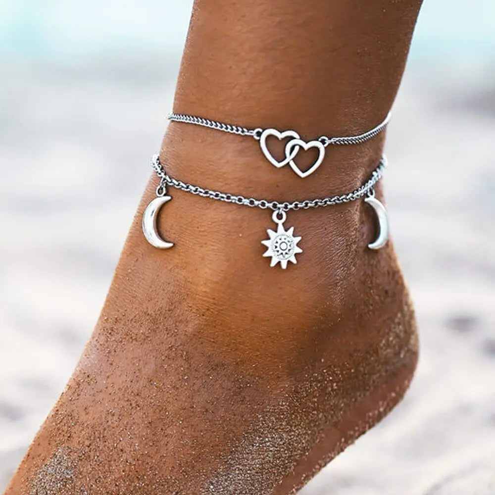 Silver anklets with heart, moon, and sun charms on a person's ankle against a blurred beach background.