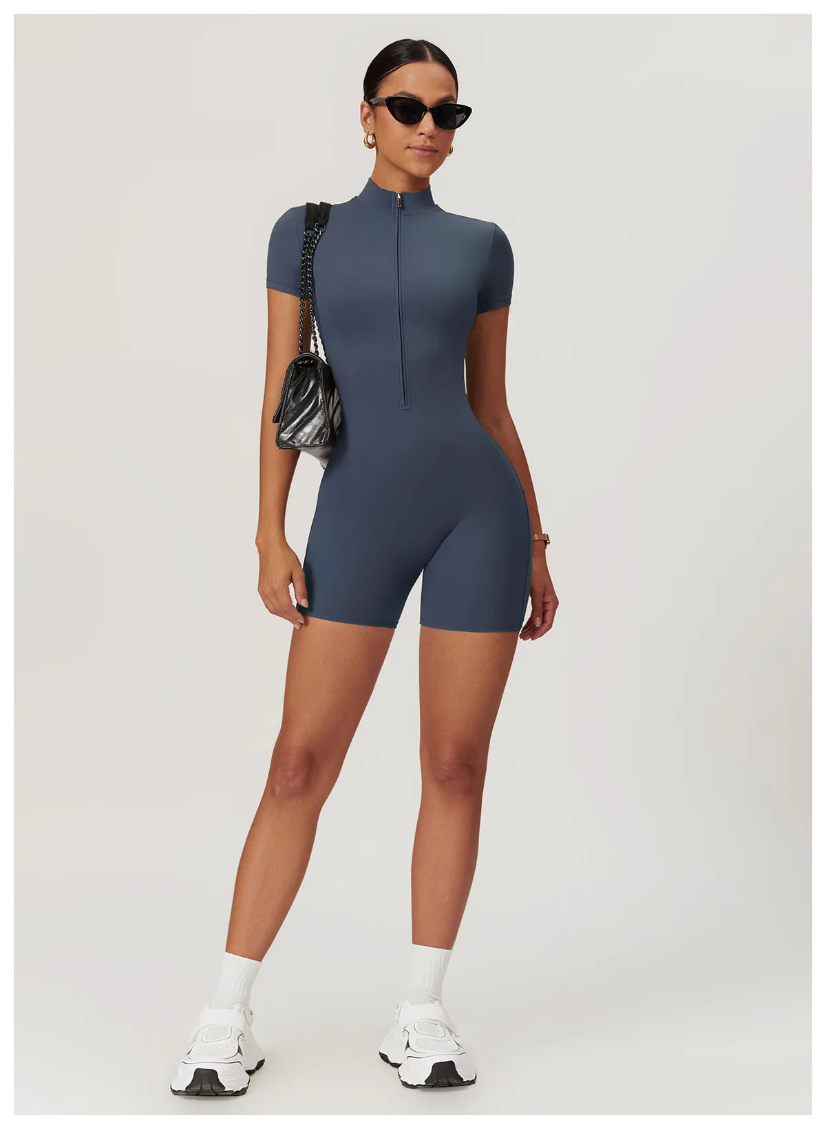 Woman wearing a blue bodysuit with sunglasses and a black handbag on a white background