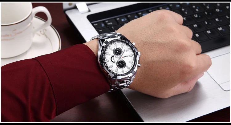 Hand wearing a silver watch with a white face on a desk with a laptop and cup.
