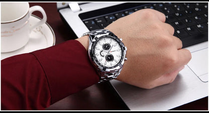 Hand wearing a silver watch with a white face on a desk with a laptop and cup.