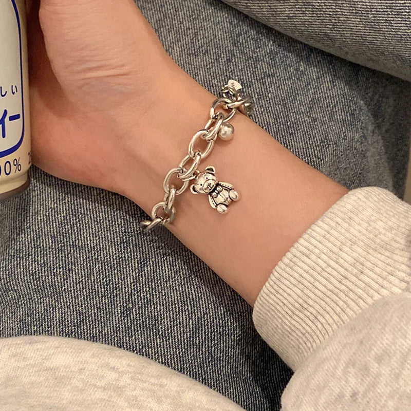 Silver chain bracelet with a teddy bear charm on a wrist, against a gray fabric background.