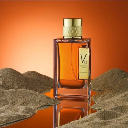 Perfume bottle with gold cap and label on a reflective surface with sand-like texture and gradient orange background