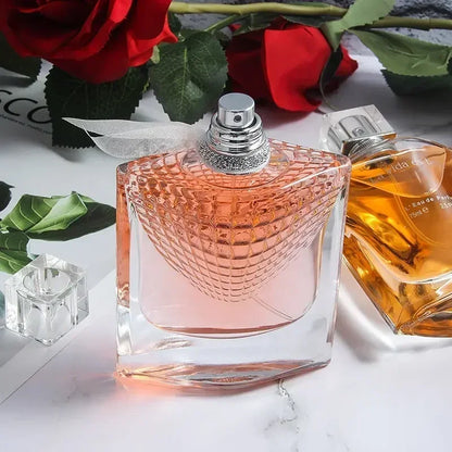 Pink perfume bottle with textured design on a marble surface with red roses and another perfume bottle in the background.