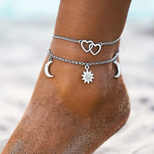 Silver anklets with heart, moon, and sun charms on a person's ankle against a blurred beach background.