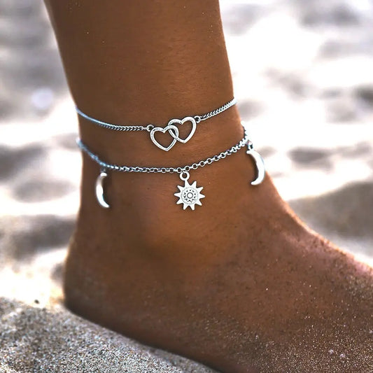 Silver anklets with heart, moon, and sun charms on a foot against a blurred beach background