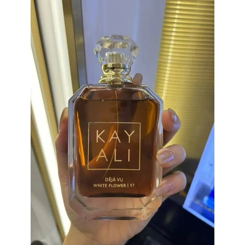 Hand holding a Kayali perfume bottle labeled 'Deja Vu White Flower 57' against a blurred background.