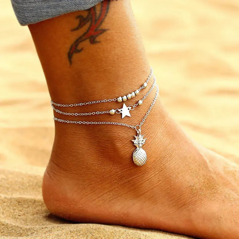 Ankle with multiple silver anklets, including a pineapple charm, on a sandy background.